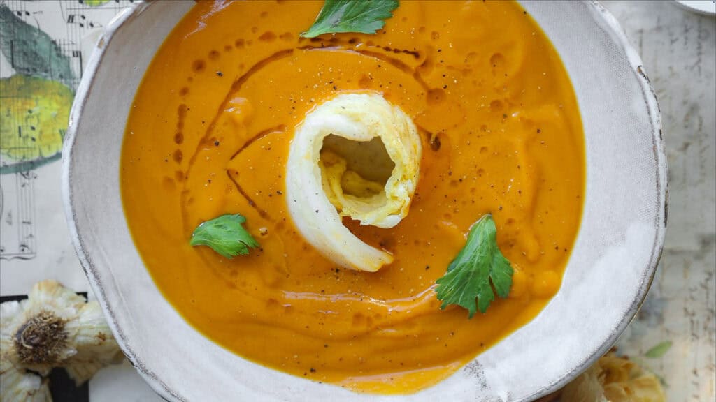 A bowl of gochujang and roasted carrot soup, garnished with a swirl of cream and fresh parsley leaves. The soup is in a white bowl placed on a patterned surface with hints of garlic visible nearby.